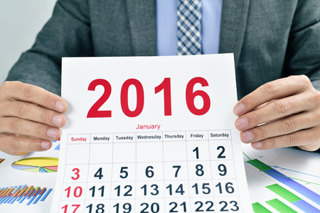 a young caucasian businessman at his office desk full of charts shows a 2016 calendarの写真素材