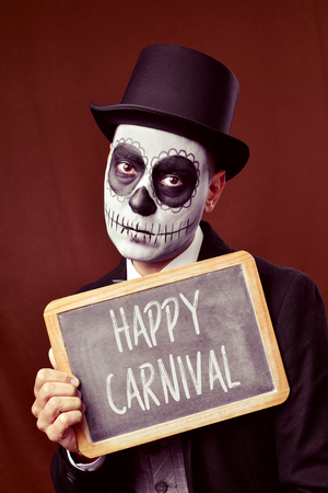 a man with mexican calaveras makeup, wearing jacket, vest, bow tie and top hat, shows a chalkboard with the text happy carnivalの写真素材
