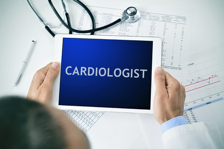 closeup of a doctor man sitting at his office desk holding a tablet computer with the word cardiologist in its screenの写真素材