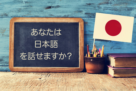 a chalkboard with the question do you speak Japanese? written in Japanese, a pot with pencils, some books and the flag of Japan, on a wooden deskの写真素材