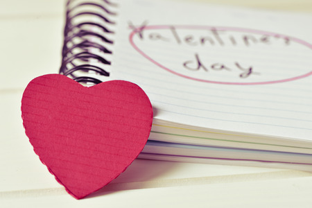 closeup of a red hart and a notebook with the text valentines day handwritten in it, on a white surface, with a filter effectの写真素材