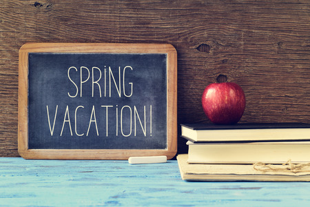an old chalkboard with the text spring vacation written in it, a piece of chalk, and a red apple on a pile of books, on a blue rustic wooden tableの写真素材