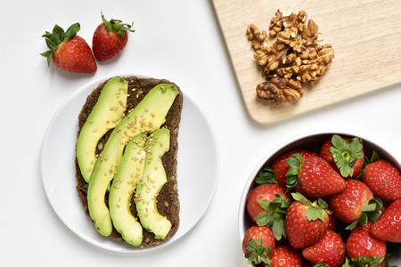 high-angle shot of an avocado toast in a white plate, a white bowl full of strawberries and some peeled walnuts on a wooden chopping board, on a white surfaceの写真素材