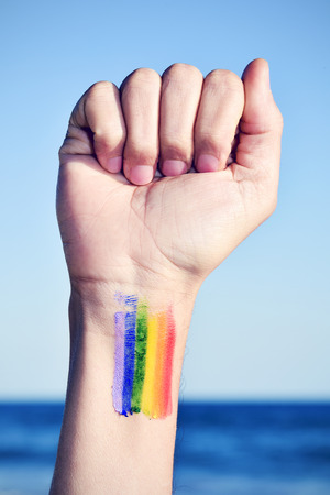 closeup of a young caucasian man with his fist raised to the sky and a rainbow flag painted in his wrist, with the ocean in the backgroundの写真素材
