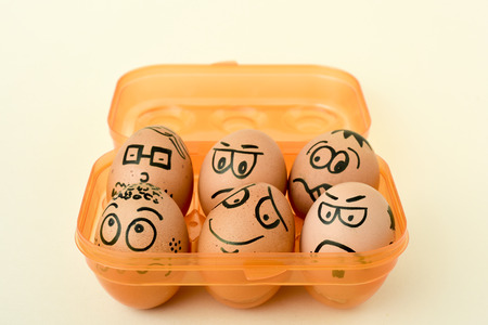 a pile of brown eggs ornamented with funny faces in an orange egg box, on an off-white backgroundの写真素材