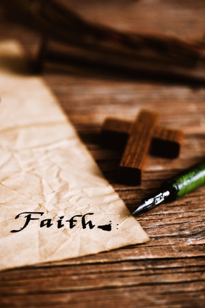 closeup of a small wooden christian cross, an old dip pen and a yellowish paper with the word faith written with black ink in it, on a rustic wooden tableの写真素材