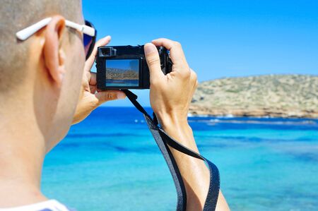 closeup of a young caucasian man seen from behind taking a picture of the Mediterranean sea and its coast in Ibiza Island, Spain, in the summerの写真素材