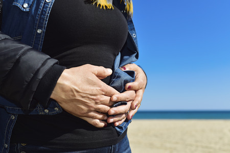 closeup of the hands of a young man on the hands of a young woman in her belly whom he is embracing from behind, in front of the seaの写真素材