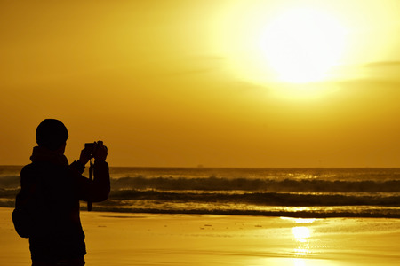 the silhouette of a young caucasian man wearing scarf and knit cap taking a picture in front of the sea at dusk, against a colorful orange skyの写真素材