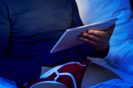 a young caucasian man lying in bed has his hand under his underwear while is watching his tablet