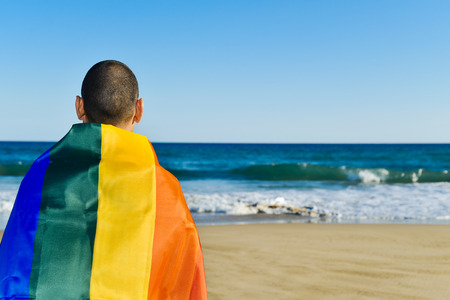 closeup of a young caucasian man seen from behind wrapped in a rainbow flag looking at the oceanの写真素材