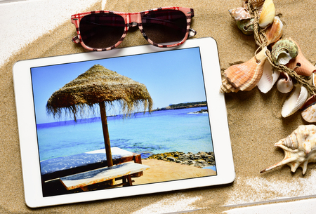 high-angle shot of a white wooden surface full of sand and an ornament made with different conchs and seashells, a pair of sunglasses and a tablet computer with the picture of a beach taken by myselfの写真素材