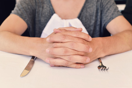 closeup of a young caucasian man sitting at a table waiting for the food, with his hands clasped a and a knife and a fork in front of himの写真素材