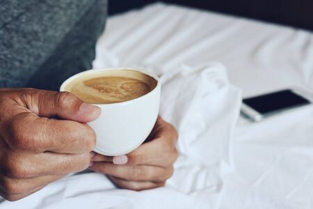 closeup of a young caucasian man in pajamas with a cup of coffee in his hand in bed, and a smartphone on the white sheet in the backgroundの写真素材
