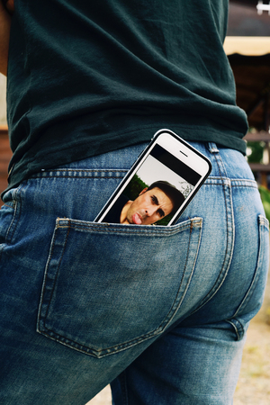 a smartphone with a picture of a young man sticking his tongue out in its screen, taken by myself, placed on the back pocket of the jeans of a young manの写真素材