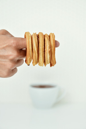 closeup of the hand of a young man with some Spanish churros in his forefinger and a cup of hot chocolate in the backgroundの写真素材