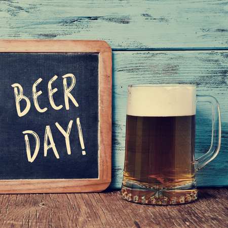 an old chalkboard with the text beer day written in it and a jar with refreshing beer on a rustic wooden table, against a blue wooden backgroundの写真素材