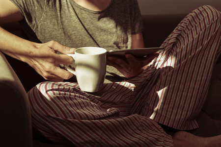 closeup of a young caucasian man in pajamas with a cup of coffee in his hand using a tablet computer seating in a couch, illuminated by some sunbeamsの写真素材