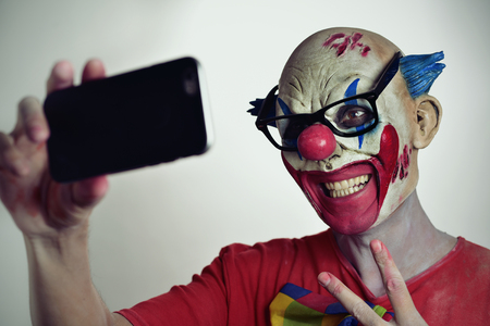 portrait of a scary evil clown taking a selfie with his smartphone, while giving a V signの写真素材