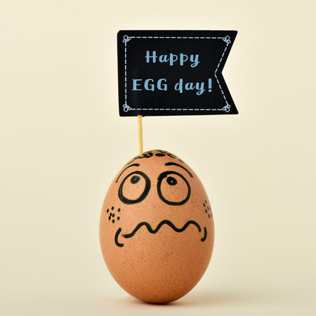 a brown egg with a funny face with a black flag-shaped signboard with the text happy egg day, on an off-white backgroundの写真素材