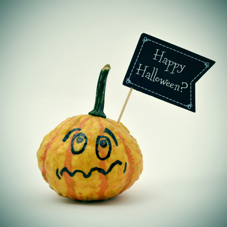 closeup of a terrified pumpkin with a black flag-shaped signboard with the question Happy Halloween? written in it, against an off-white backgroundの写真素材