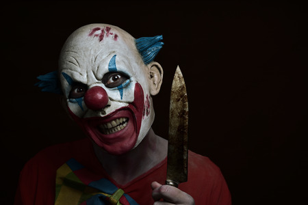 a scary evil clown with a big knife in his hand, against a dark backgroundの写真素材
