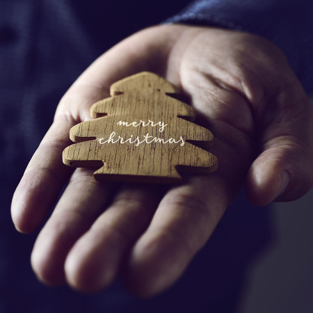 closeup of a wooden christmas tree with the text merry christmas written in it, in the palm of the hand of a young caucasian manの写真素材