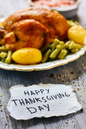 a piece of paper with the text happy thanksgiving day handwritten in it and a roast turkey on a round ceramic tray, placed on a rustic wooden tableの写真素材