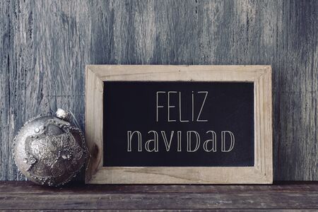 a wooden-framed chalkboard with the text feliz navidad, merry christmas written in spanish, and a christmas ball, on a rustic wooden surfaceの写真素材