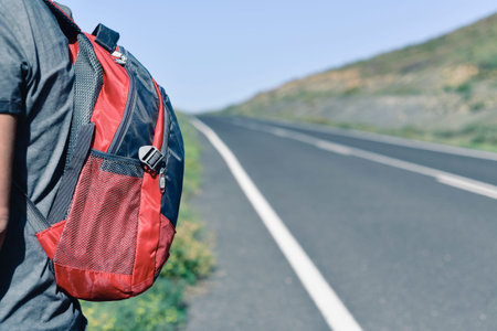 closeup of a young caucasian man seen from behind carrying a backpack standing next to a minor road