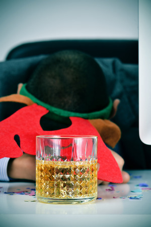 closeup of a young caucasian businessman wearing a reindeer antler headband sleeping at his desk, covered with confetti and with a glass of liquor in the foreground, after an office christmas partyの写真素材