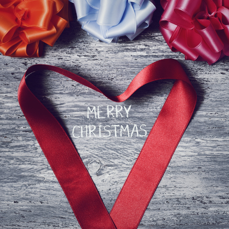 some satin gift ribbon bows of different colors and a red satin ribbon forming a heart on a rustic wooden surface and the text merry christmas in the centerの写真素材