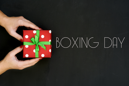 high-angle shot of a young caucasian woman with a gift in her hands wrapped in a red paper patterned with white dots and tied with a green ribbon, and text boxing day written on a dark gray surfaceの写真素材