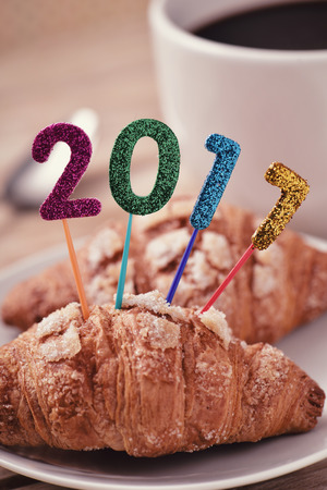 closeup of four glittering numbers of different colors forming the number 2017, as the new year, topping a small croissant and a cup of coffee placed on a table set for breakfastの写真素材