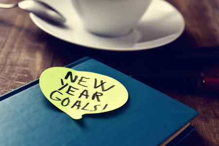 a yellow sticky note in the shape of a speech balloon with the text new years goals written in it attached to a black notepad on a rustic wooden table next to a cup of coffeeの写真素材