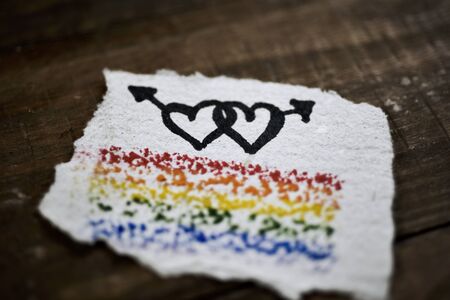 closeup of a piece of paper with a rainbow flag and two hearts in the shape of the male gender symbol painted in it, on a rustic wooden surfaceの写真素材