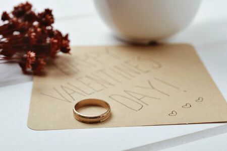 closeup of a golden ring and a brown paper note with the text happy valentines day written in it, on a white table next to a bunch of dry flowersの写真素材