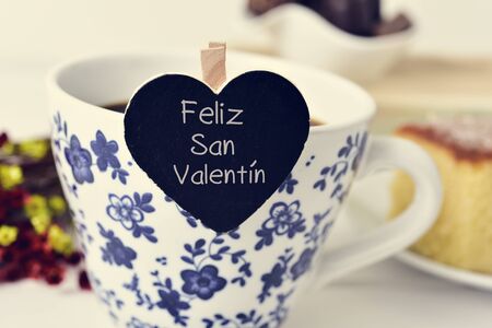 closeup of a heart-shaped signboard with the text feliz san valentin, happy valentines day written in spanish, in a cup of coffee, on a table set for breakfastの写真素材
