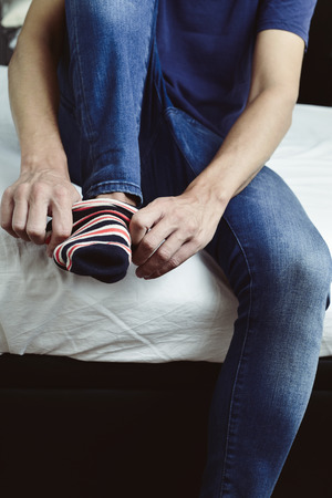 closeup of a young caucasian man wearing sitting on the edge of the bed putting on or taking off a pair of colorful striped socksの写真素材
