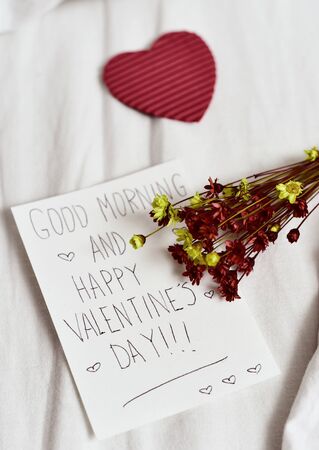 closeup of a white paper note with the text good morning and happy valentines day written in it, a red heart and a bunch of dried flowers, on the white sheets of an undone bedの写真素材