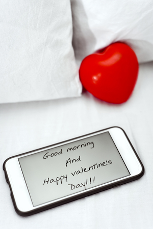 a smartphone, with the text good morning and happy valentines day in its screen, and a red heart placed on the white sheets of a bedの写真素材