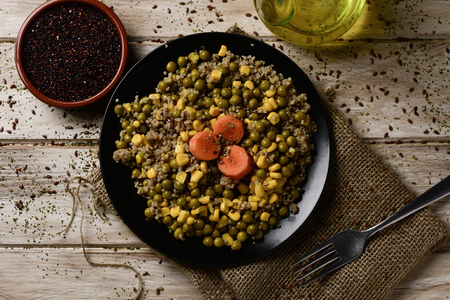 high-angle shot of a black ceramic plate with a salad made with quinoa, sweet corn, green peas and carrot, placed on a rustic wooden table next to a cruet with olive oil and a bowl with quinoa seedsの写真素材