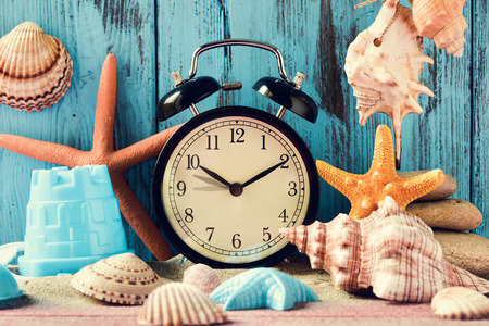 an alarm clock being adjusted forward one hour at the beginning of the summer time, many conches and starfishes, and a beach pail, on a pile of sand, against a bright blue rustic wooden backgroundの写真素材