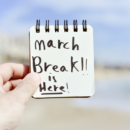 closeup of a the hand of a young caucasian man holding a notepad with the text spring break is here written in it, in front of a beachの写真素材