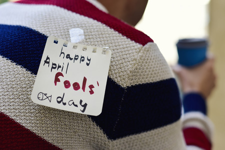 closeup of a young caucasian man seen from behind having a coffee, with a note with the text happy april fools day attached with tape to his backの写真素材