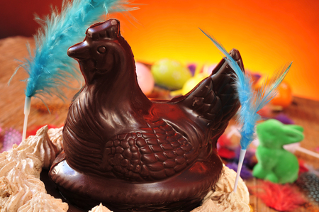 closeup of a mona de pascua, a cake eaten in Spain on Easter Monday, topped with a chocolate chicken, on a rustic wooden surface full of decorated eggs and feathers of different colorsの写真素材