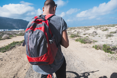 a young man with a backpack seen from on a bike, stopped to look to his smartphone or GPS, in a dirty road in La Graciosa, Canary Islands, Spainの写真素材