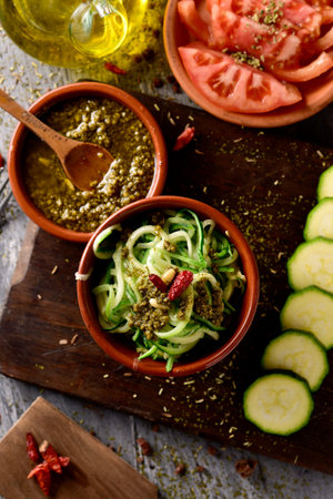 high-angle shot of some earthenware bowls with zucchini spaghetti, italian pesto sauce and chopped tomato, and a cruet with olive oil on a rustic wooden tableの写真素材