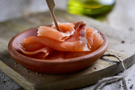 closeup of an earthenware bowl with some slices of smoked salmon, on a rustic wooden chopping board and a cruet of olive oil in the backgroundの写真素材