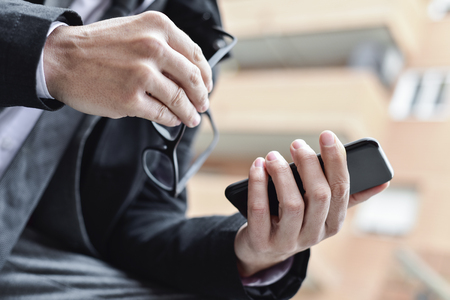 closeup of a young caucasian businessman in a gray suit outdoors using a smartphone with one hand and holding his eyeglasses in the other handの写真素材
